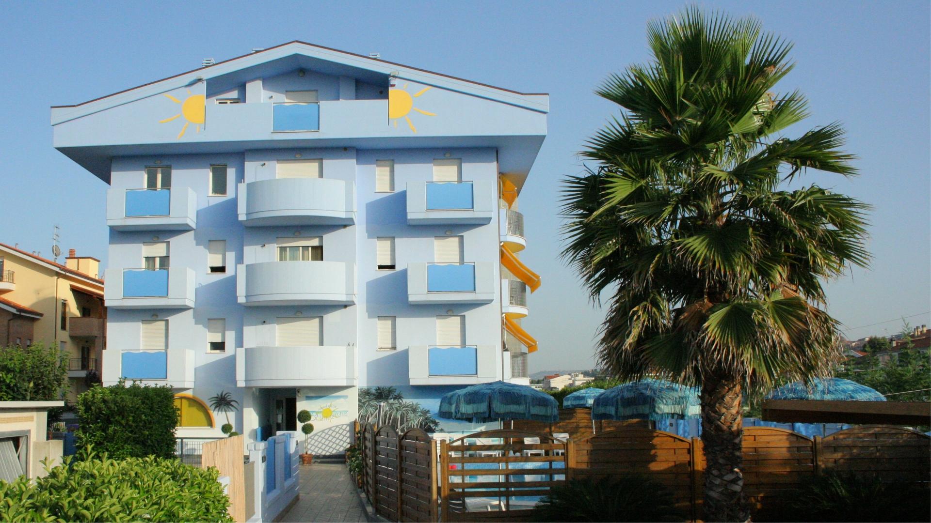 Edificio azzurro con balconi, piscina e palme, sotto un cielo sereno.