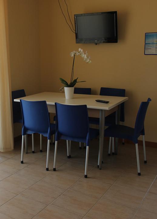 Sala da pranzo con tavolo bianco, sedie blu, TV a parete e pianta ornamentale.